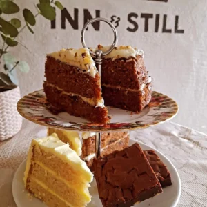 An assortment of delicious cakes, including slices of carrot cake, lemon cake, and brownies, displayed on a floral two-tiered cake stand at Inn & Still in Helston, Cornwall, offering a tempting treat for visitors.