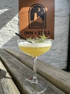 Elegant cocktail garnished with rosemary served in a crystal glass, placed on a rustic wooden bench in front of the Inn & Still sign, highlighting the stylish and relaxed outdoor setting of the bar in Helston, Cornwall.