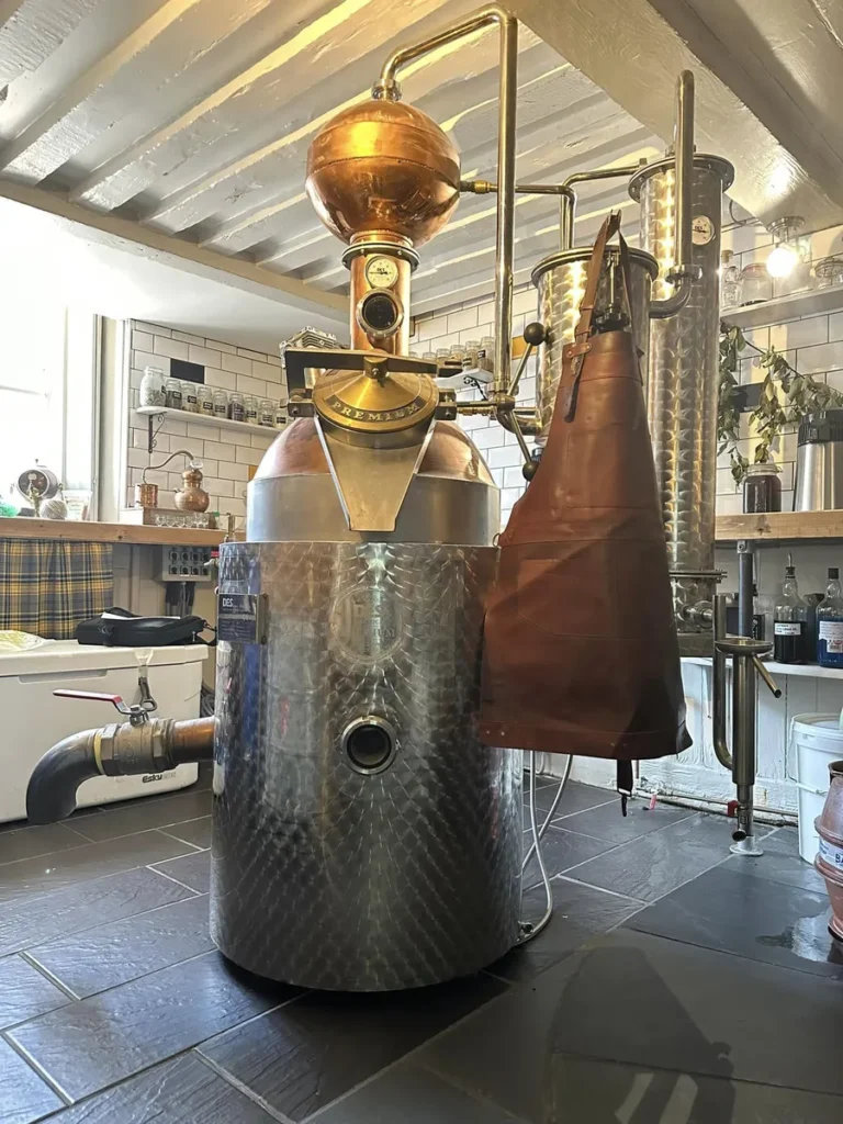 Close-up of a copper and stainless steel distillation still used for making gin, vodka, and rum at Inn & Still in Helston, Cornwall, set in a rustic room with exposed beams and a tiled floor, showcasing the traditional craftsmanship involved in spirit production.