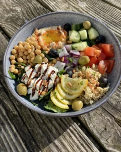 Fresh and colourful Mediterranean-style salad served in a grey bowl on a rustic wooden table, featuring a mix of chickpeas, feta cheese drizzled with balsamic glaze, hummus, couscous, olives, cucumber, tomatoes, red onion, and lemon slices