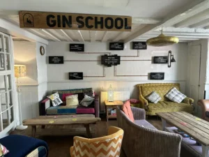 Bright and inviting seating area at Inn & Still in Helston, Cornwall, with a 'Gin School' sign above, featuring eclectic vintage furniture, cosy cushions, and a rustic wooden table, creating a comfortable and stylish atmosphere.