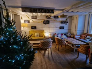 Cosy interior of Inn & Still in Helston, Cornwall, decorated for the festive season with a Christmas tree adorned with blue lights, garlands, and a 'Gin School' sign above a seating area with vintage furniture, creating a warm and inviting holiday atmosphere.