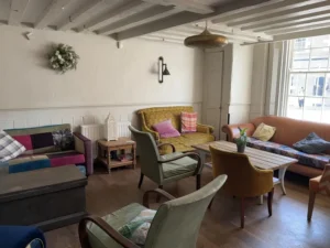 Cosy seating area at Inn & Still in Helston, Cornwall, featuring a mix of vintage sofas and armchairs with colourful cushions, set in a bright room with wooden beams and natural light streaming through a large window.