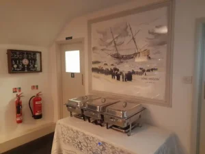 pstairs Anson Bar at Inn & Still in Helston, featuring a buffet setup with chafing dishes on a lace-covered table beneath a large framed painting of HMS Anson, highlighting the bar’s nautical theme and historical connection.