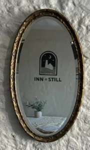 Ornate oval mirror reflecting the Inn & Still logo, mounted on a textured stone wall, with a small potted plant visible in the reflection, adding a touch of elegance to the rustic decor.