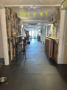 Interior hallway of Inn & Still in Helston, Cornwall, featuring rustic decor with exposed wooden beams, bunting, and fairy lights, leading to a bright seating area in the distance, creating a warm and inviting atmosphere.