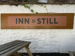 Outdoor sign for Inn & Still in Helston, Cornwall, displayed on a whitewashed stone wall above a rustic wooden bench, giving a glimpse of the establishment's charming and relaxed atmosphere.