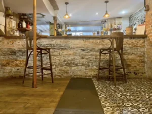 Rustic bar area at Inn & Still in Helston, Cornwall, featuring a weathered wooden bar front, vintage high stools, and patterned floor tiles, creating a cosy and inviting space