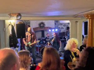 Live performance by Clusterfunk band at Inn and Still, Helston, with the lead singer and guitarist engaging the audience in a cosy, intimate setting filled with lively patrons.