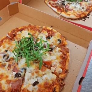 Close-up of two freshly made pizzas topped with rocket, olives, and various cheeses, served in takeaway boxes, ready to be enjoyed.