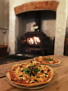 Delicious pizzas served on a rustic wooden table in front of a warm, glowing wood-burning stove at Inn and Still, Helston, Cornwall, creating a cosy and inviting dining experience.