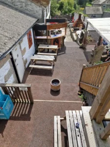 View of the pub garden at Inn and Still in Helston, Cornwall, featuring the SAR outside bar, picnic-style wooden benches, a central fire pit, and a mix of rustic and modern outdoor décor, all set within a charming courtyard area.
