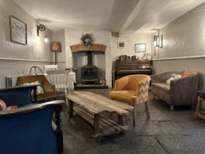 Cozy interior of Inn and Still in Helston, Cornwall, showcasing a wood-burning stove with a rustic brick arch, eclectic seating including vintage armchairs and a patterned yellow chair, a wooden coffee table, and a piano, all illuminated by warm ambient lighting to create a welcoming atmosphere for relaxation.